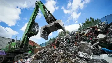 green digger outside in a recycling centre with open grabber about to pick up electricals from a mound of electrical items