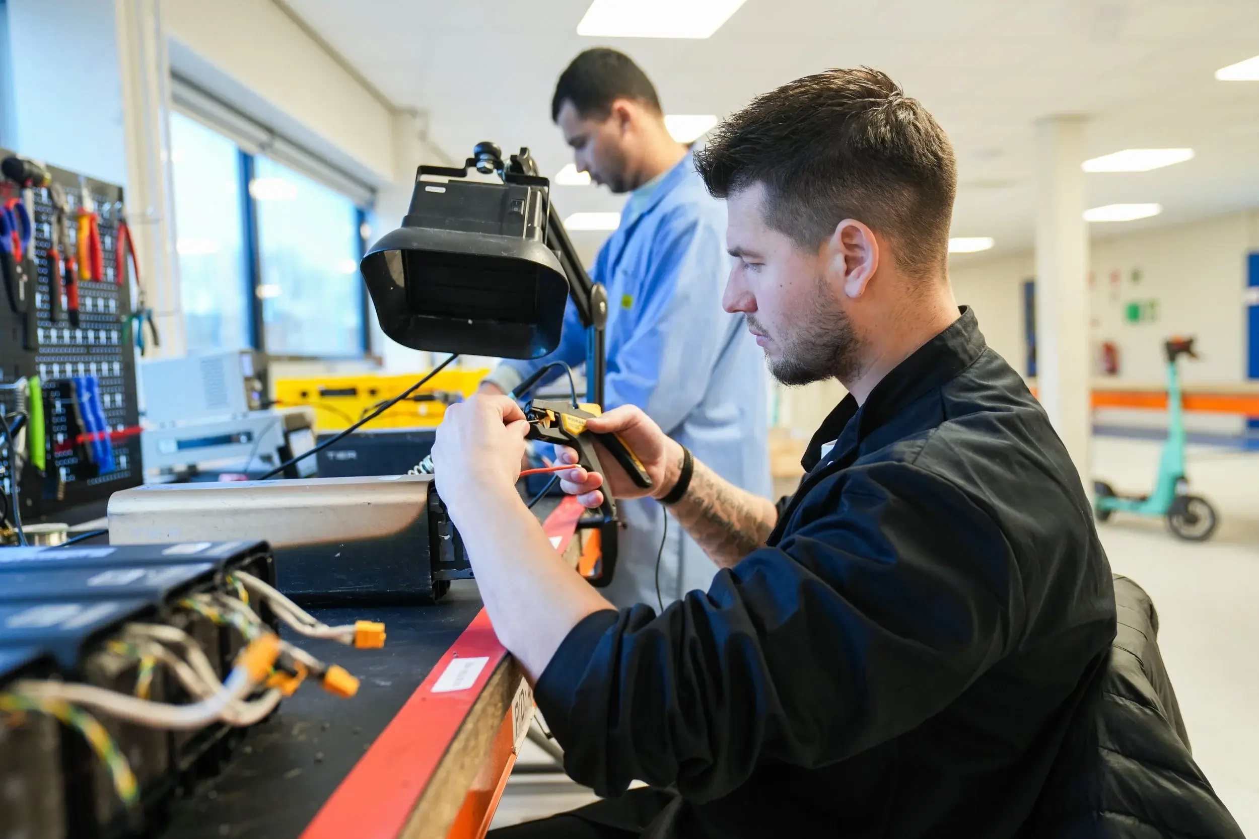 Two men working with electronic devices and tools at a workbench in a workshop or lab, with a scooter visible in the background.