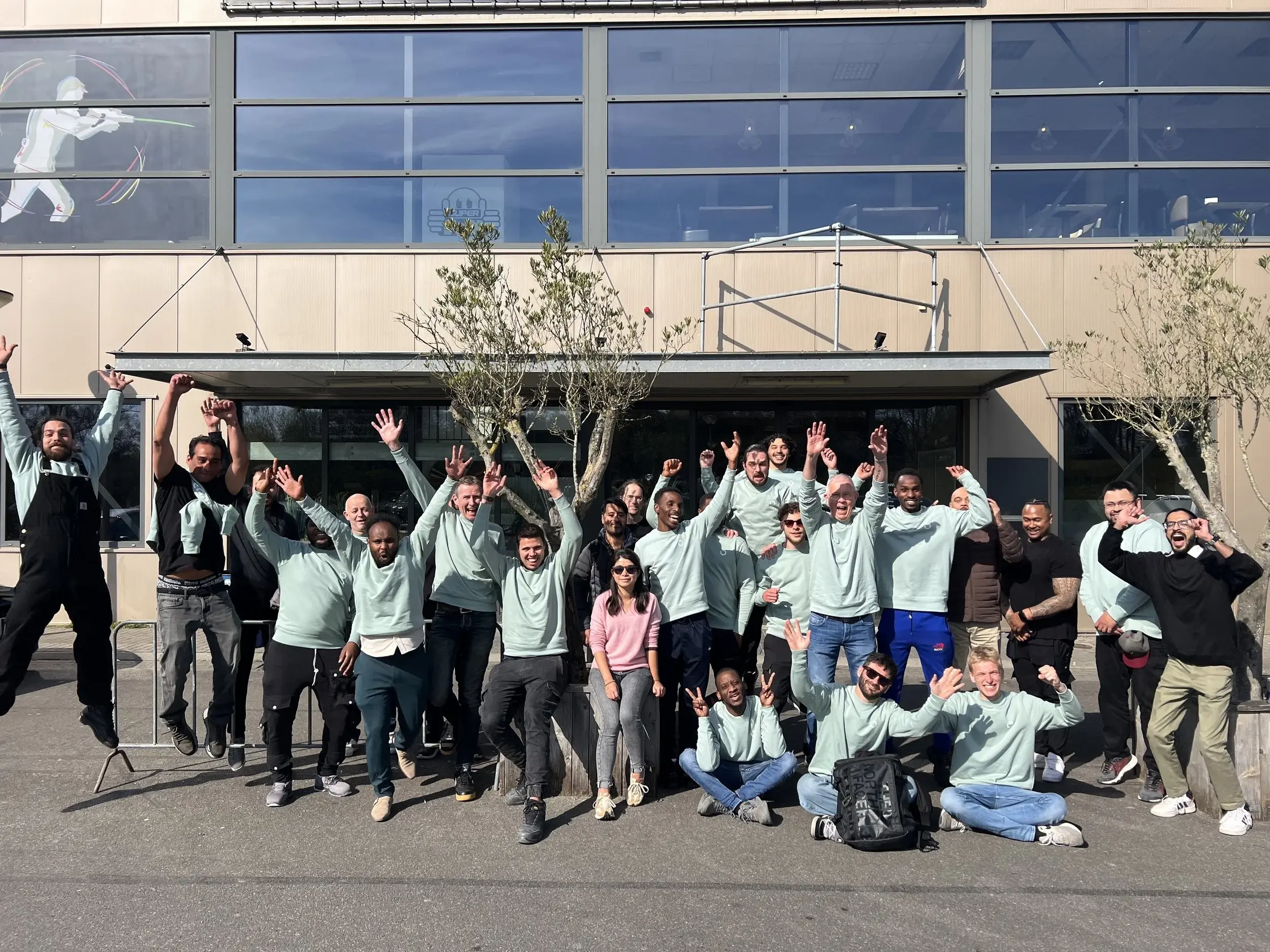 A group of people posing cheerfully outside a modern building with large glass windows, some standing, others sitting or jumping, with a few trees and clear sky in the background.